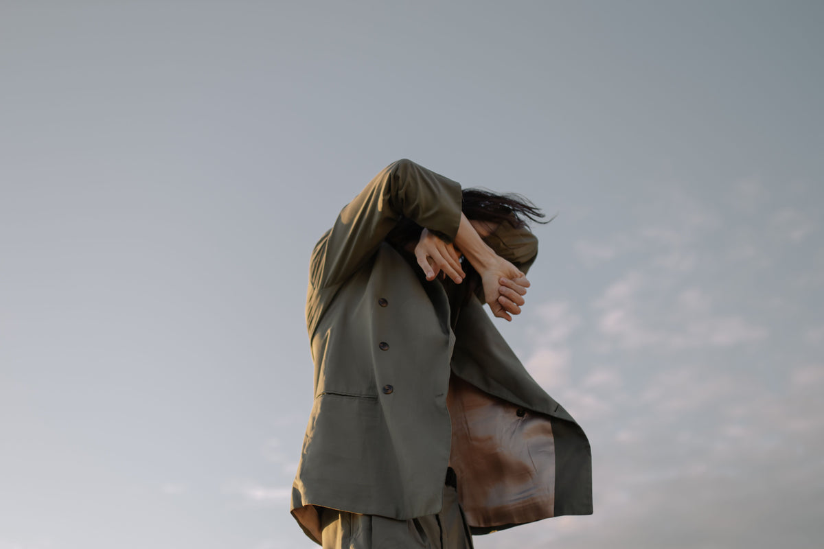 A person outdoors in a light olive green blazer and matching trousers, holding their head in their hands against a pale sky.