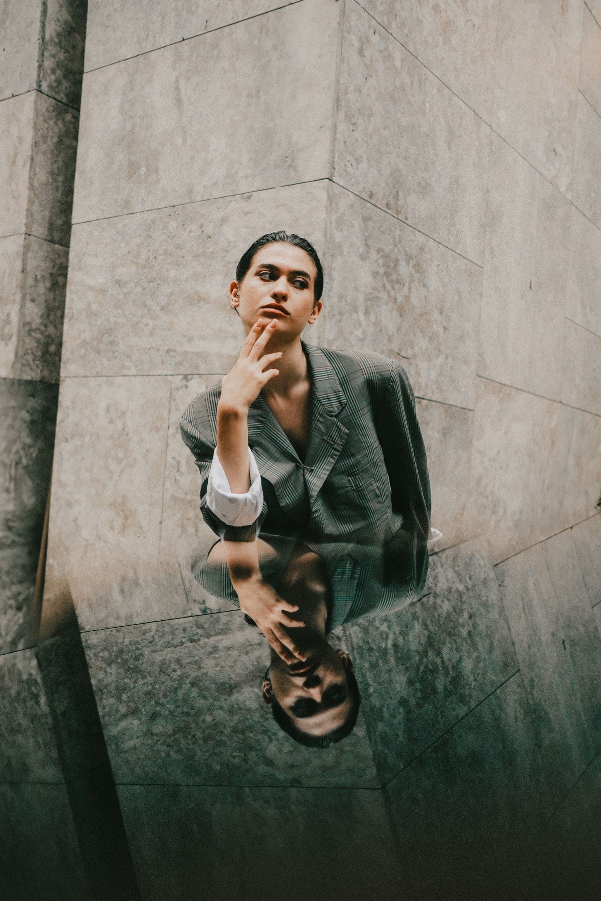 A woman in a dark gray blazer and white shirt poses in a stone-walled corridor, with her reflection visible in the wet pavement below.