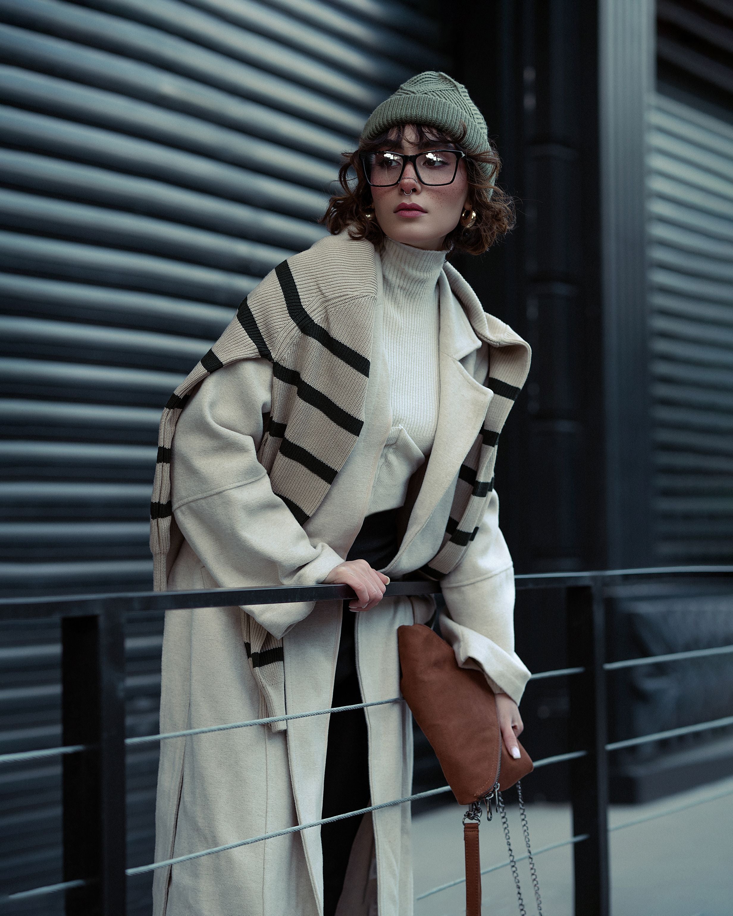 A woman wearing a long cream coat with bold black stripes, a cream turtleneck, a green knit beanie, and glasses, holding a brown leather bag while leaning on a railing in front of a dark corrugated metal wall.